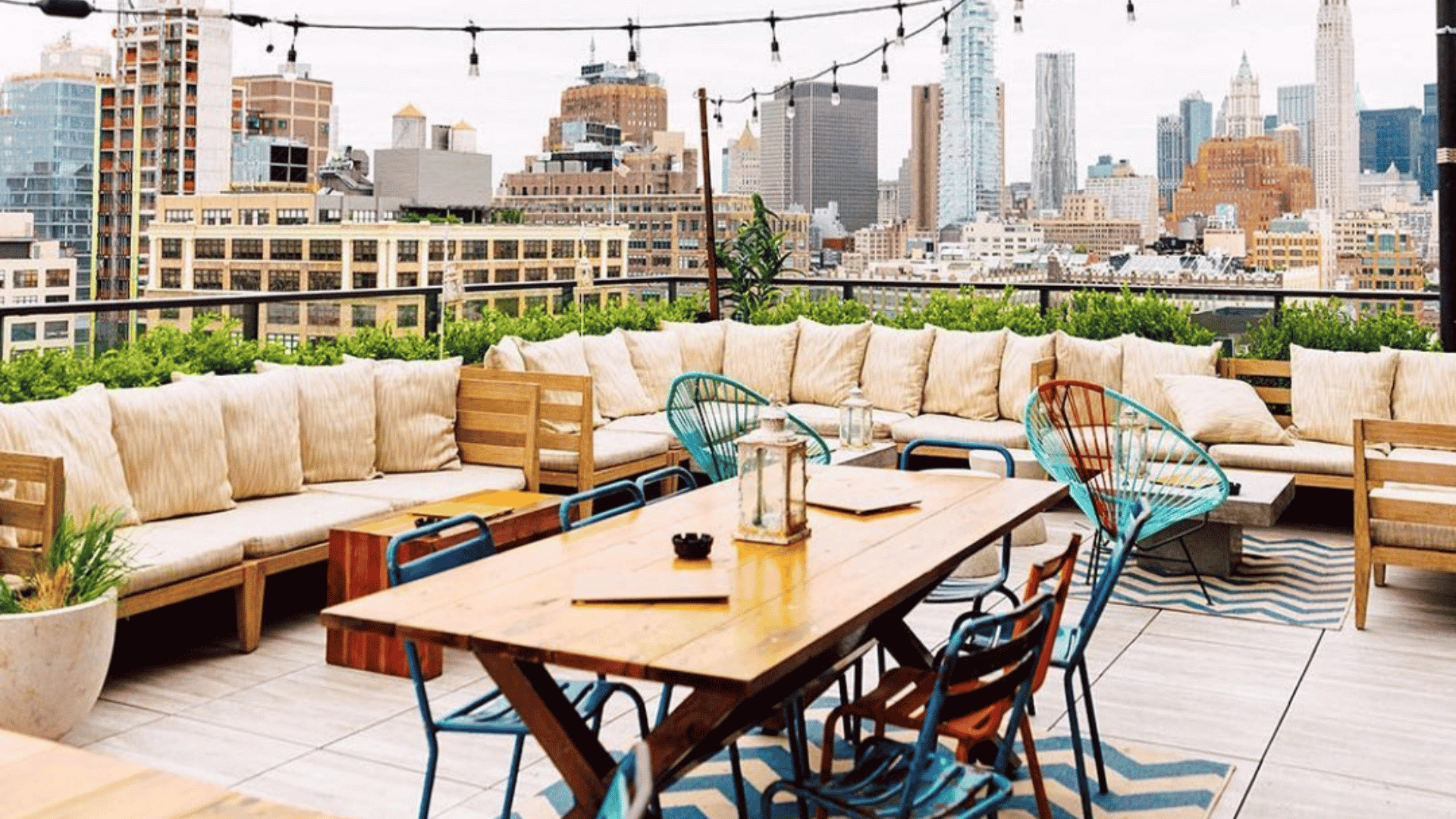 A rooftop lounge area featuring a wooden dining table, colorful metal chairs, and plush sofa seating with a view of a city skyline.