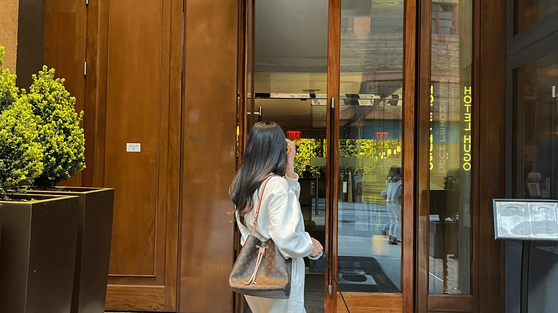A woman with long dark hair, wearing a casual white outfit and carrying a brown monogram bucket bag.