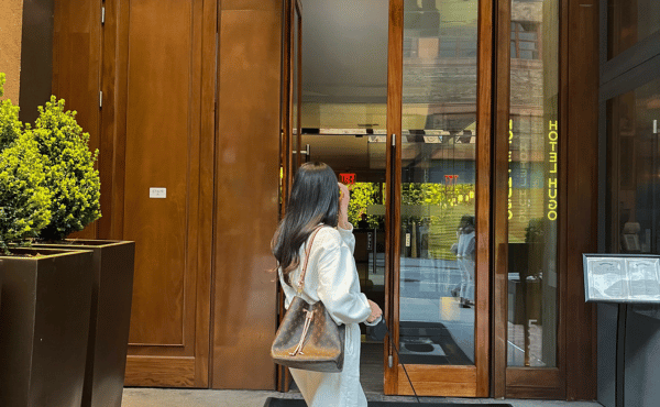 A woman with long dark hair, wearing a casual white outfit and carrying a brown monogram bucket bag.