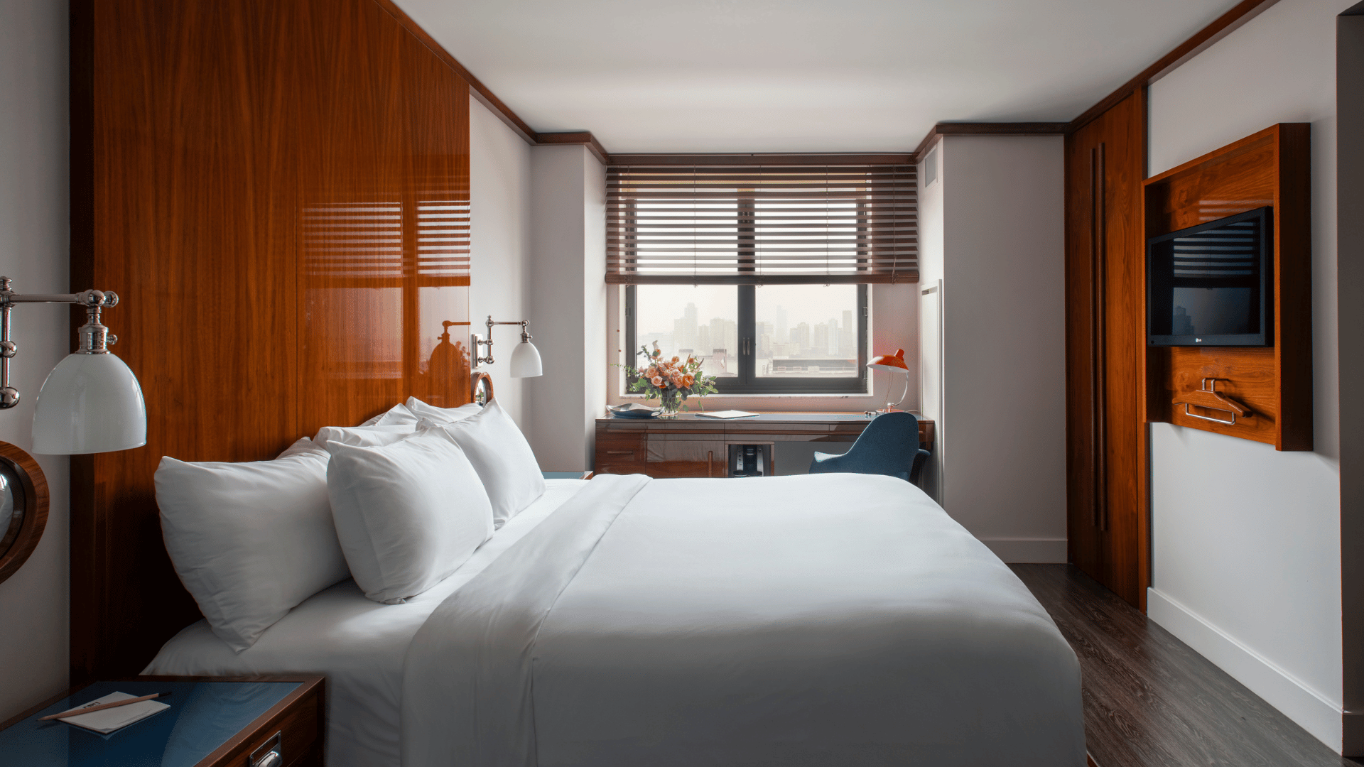 A modern hotel room featuring a large bed with white bedding, glossy wood-paneled walls, and a desk by a window overlooking a city skyline.