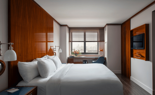 A modern hotel room featuring a large bed with white bedding, glossy wood-paneled walls, and a desk by a window overlooking a city skyline.