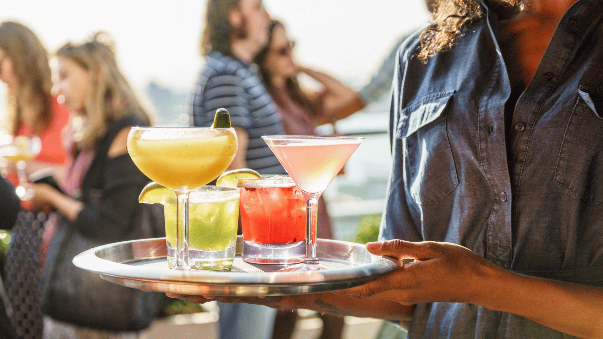 Professional server carrying a tray of handcrafted cocktails on the rooftop at Hotel Hugo NYC during a lively evening with a festive atmosphere.