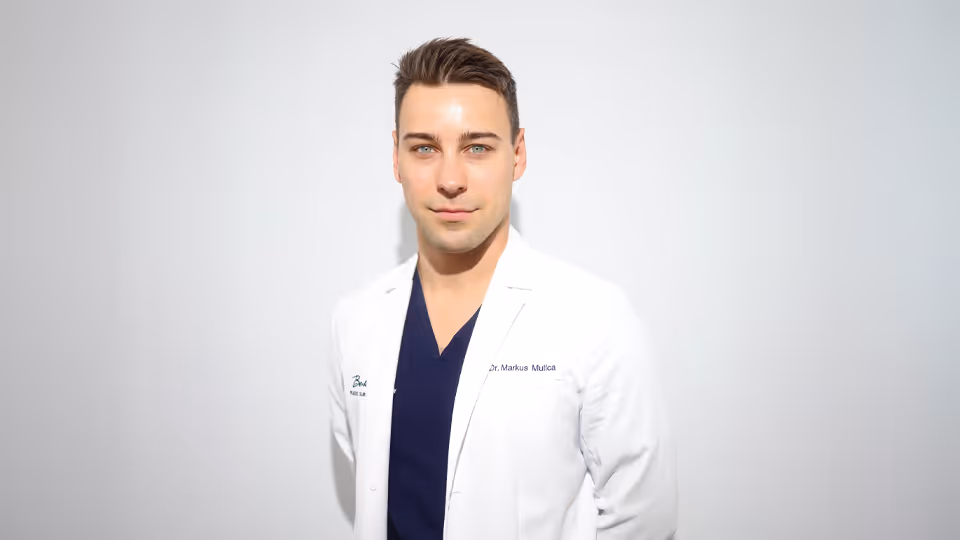 Dr. med. Markus Mulica, Facharzt für Plastische, Ästhetische und Rekonstruktive Chirurgie mit weißen Kittel und dunkelblauem Hemd vor hellgrauem Hintergrund.