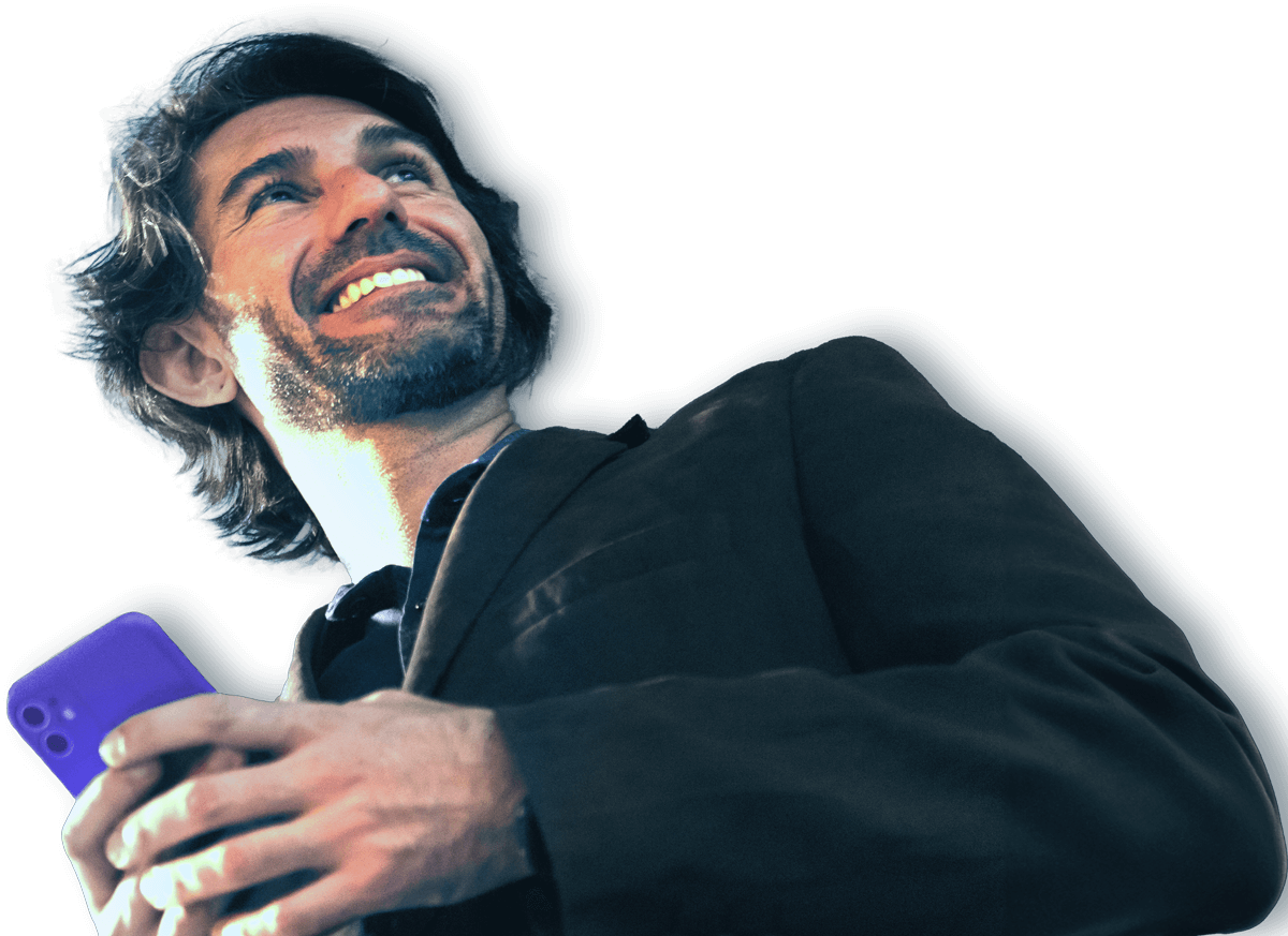 A man smiling while holding a smartphone in his right hand, wearing a dark jacket. He appears to be looking slightly upward, with light illuminating his face against a plain background.