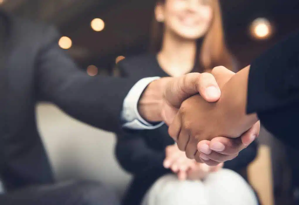 A close-up of two people shaking hands with a third person, slightly out of focus, smiling in the background. All individuals appear to be dressed in business attire, suggesting a professional or corporate setting. The interaction hints at discussions involving IDIQ services or identity theft protection strategies.