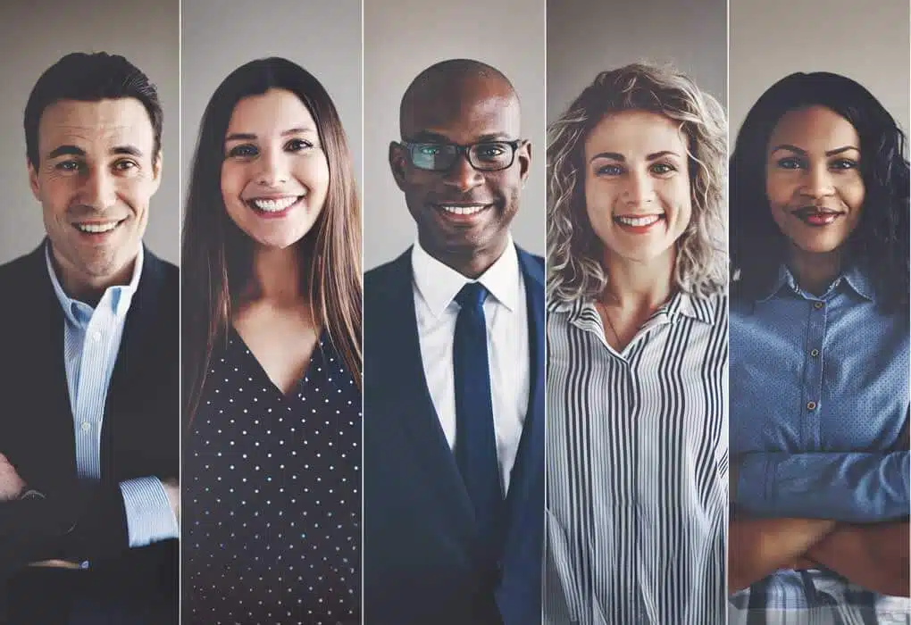 An image of a diverse group of five smiling professionals standing in a row. They are dressed in business attire and looking directly at the camera. Each individual appears confident and approachable, embodying the value of employee benefits. The background is a neutral, light-colored wall.