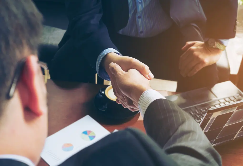 Two people in business attire are shaking hands over a wooden desk. One has glasses, and their open laptop and a document with charts are visible on the desk. A cup of coffee sits nearby, and a third person is partially visible in the background.