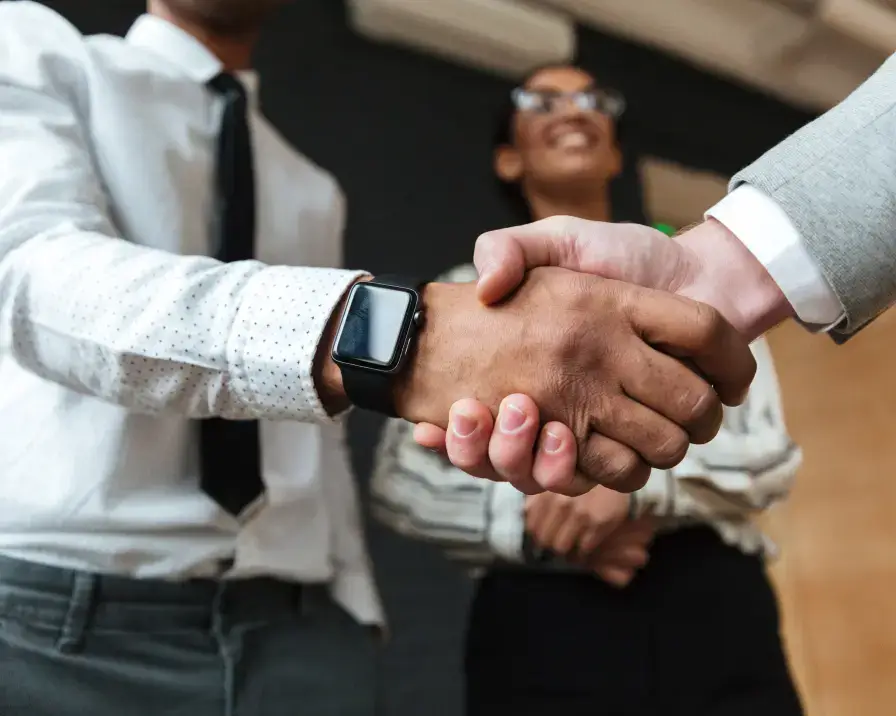 Three people in business attire are shown in a professional setting. Two individuals are in the foreground shaking hands, one wearing a smartwatch, possibly discussing identity theft protection. A third person stands in the background, slightly out of focus and smiling.