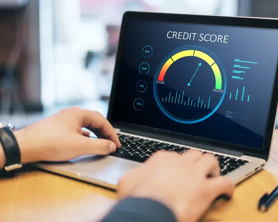 A person uses a laptop displaying a credit score dashboard. The screen shows a gauge with a needle pointing to the "Good" section and various data points. The workspace includes a desk and blurry office background, indicating a focus on credit management and financial assessment.
