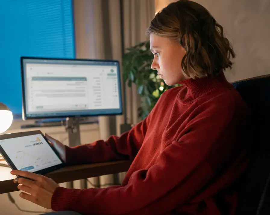 A person wearing a red sweater is sitting at a desk, looking intently at a tablet. In the background, a large computer monitor displays an employee benefits application. The room is dimly lit with a blue light coming from a nearby window.