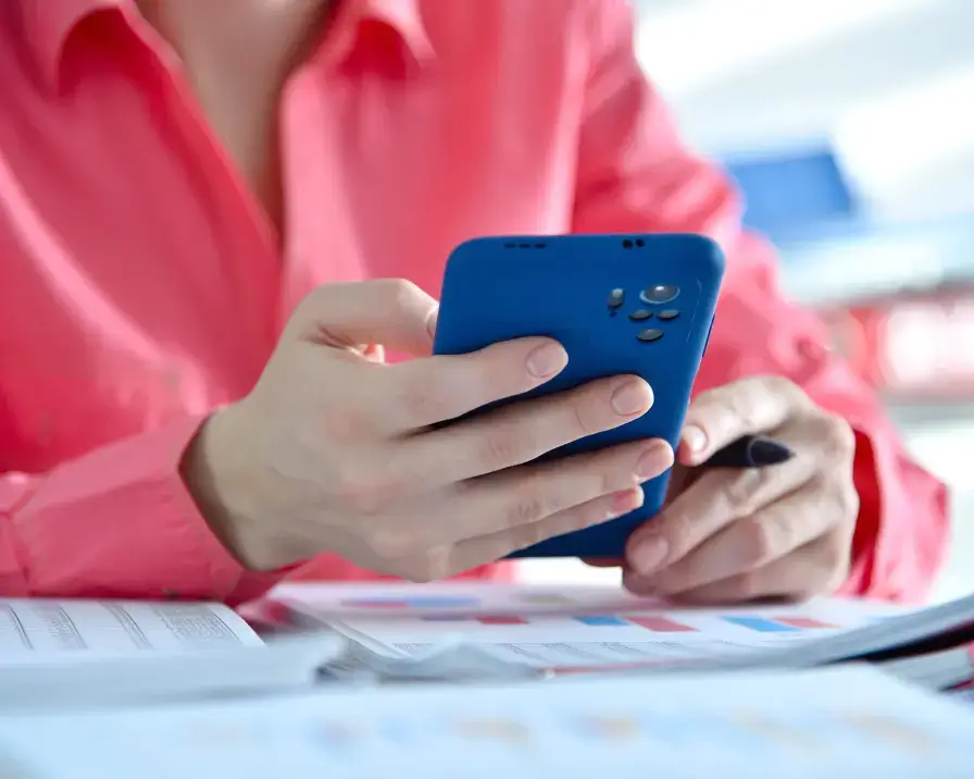 A person wearing a pink shirt is holding a blue smartphone while sitting at a desk covered in papers. The scene appears to be in a well-lit office or workspace, possibly discussing employee benefits.