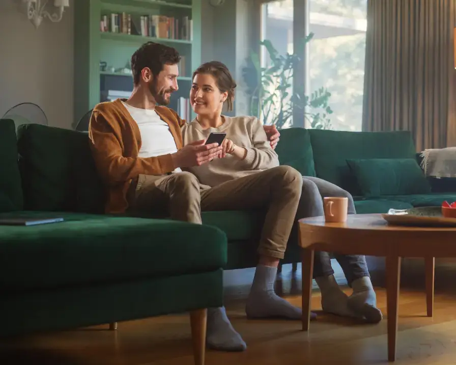 A couple sits on a green couch in a cozy living room, smiling while looking at a smartphone together. A wooden coffee table with a mug and snacks is in front of them. The room has large windows, allowing natural light to brighten the space, creating an inviting atmosphere that reflects their employee benefits plan.