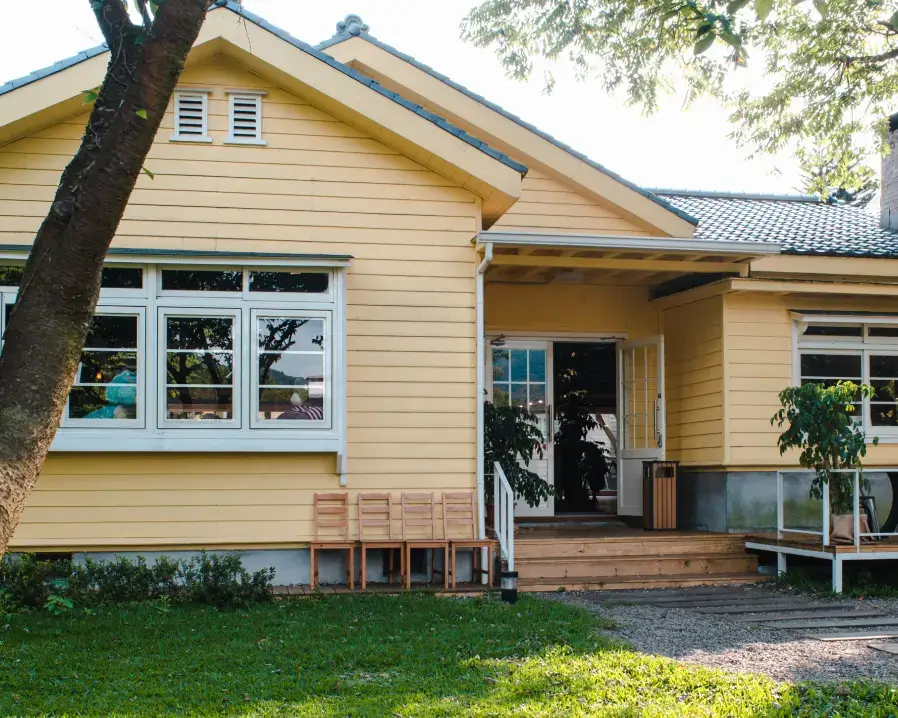 A picturesque yellow wooden house with a covered porch, white-framed windows, and a small row of wooden chairs outside. The house is set amidst a green lawn with trees casting shade. The sky is clear, indicating a sunny day—a perfect spot to unwind while considering employee benefits.