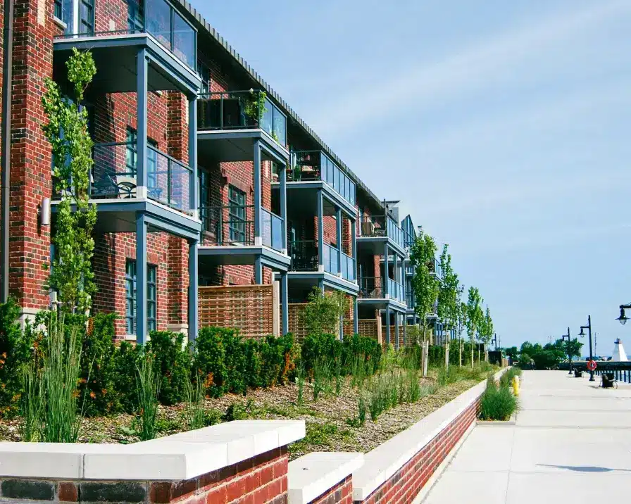 A row of modern brick apartment buildings with balconies lines a neat, tree-lined walkway. The buildings boast large windows and metal railings. Greenery, including trees and shrubs, is planted in front of the structures, adding a touch of nature to the scene while enhancing living conditions with features like rent reporting.