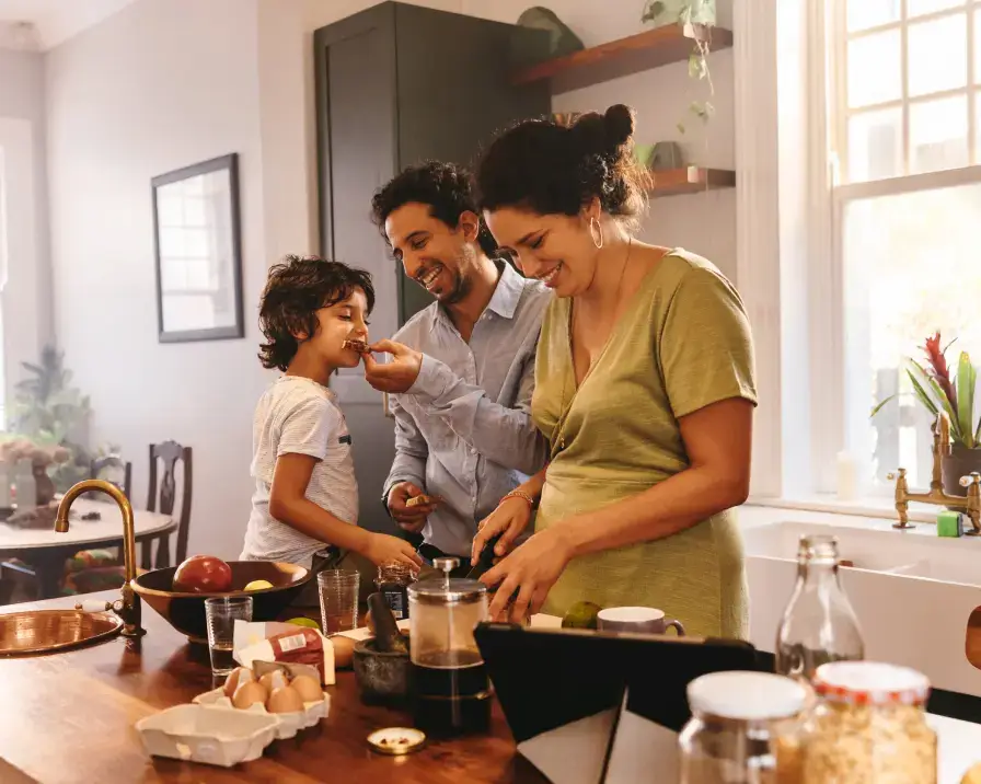 A family of three is in a cozy kitchen. The mother, wearing a green dress, is preparing food at the counter. The father, holding a piece of food, is feeding their smiling son. The kitchen features natural light, plants, and various ingredients on the counter while discussing credit management.