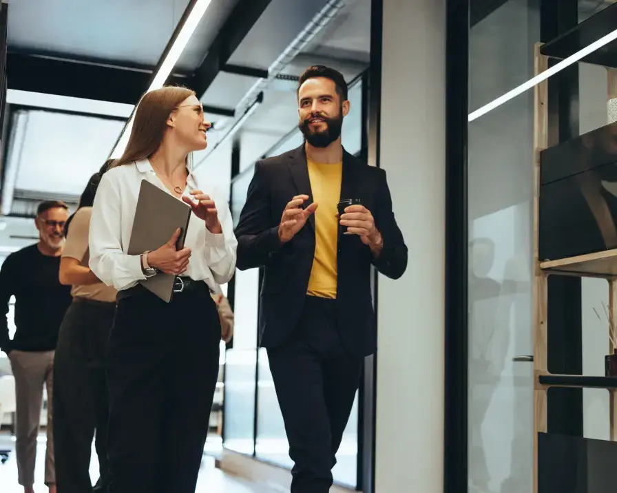 A group of colleagues is walking and chatting in an office hallway. A woman holding a laptop and a man in a suit are at the forefront, smiling and engaged in conversation about employee benefits. Other team members are visible in the background, all appearing upbeat and collegial.