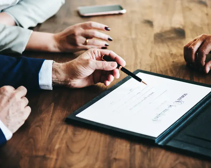Three people are sitting at a wooden table. One person is holding a black pen, pointing to a legal document on the table in a black folder. Two other people are observing. A smartphone is placed on the table in the background, possibly indicating discussions about rent reporting and credit management.