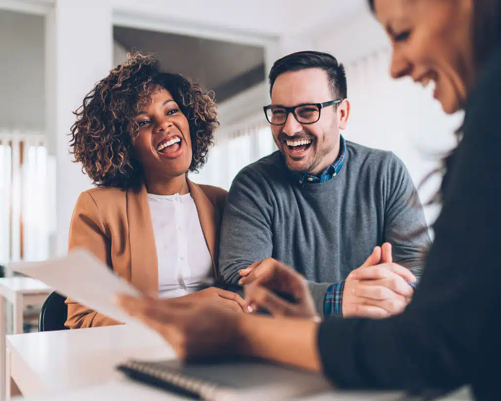 A happy couple sits together at a desk, smiling and laughing with a third person who is holding a document and a pen. The setting appears to be a professional office environment, likely discussing employee benefits or credit management.