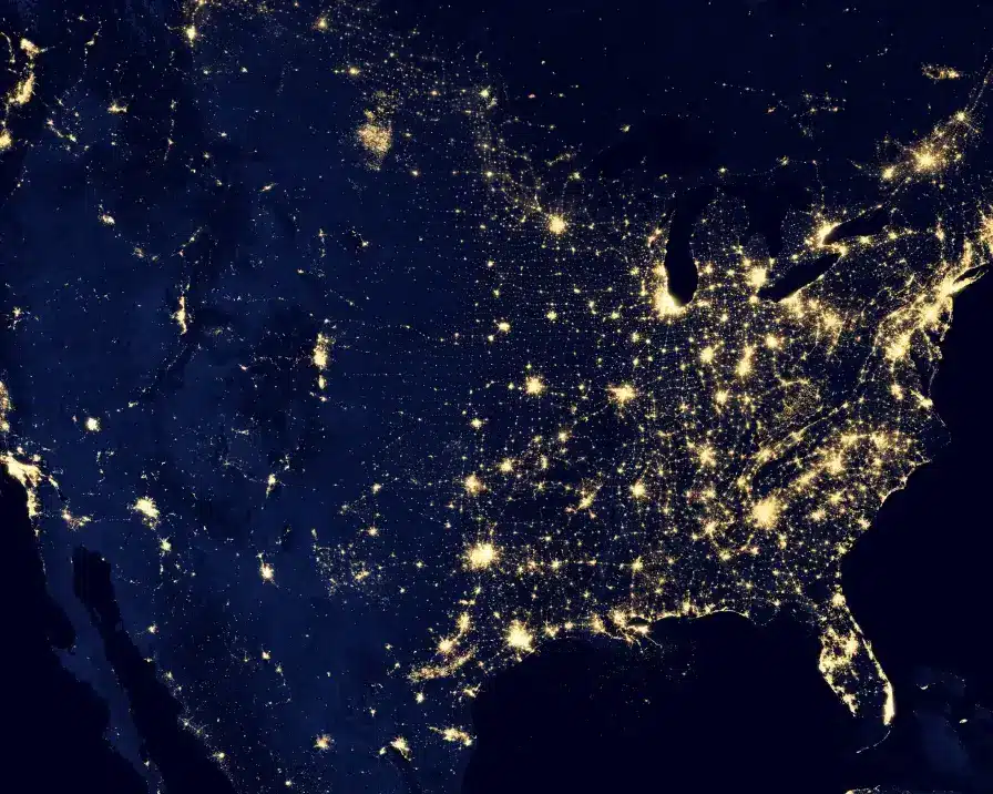 A satellite image of the United States at night, showcasing bright city lights across the eastern half of the country, particularly concentrated along the East Coast, Midwest, and Gulf Coast. The western half displays fewer and more scattered lights, indicating less urbanization. Powered by IDIQ services for identity theft protection and credit management.