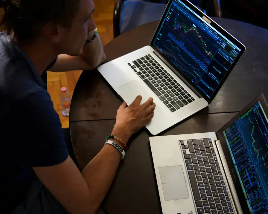 A person is seated at a round table with two laptops, focusing intently on the screens displaying complex financial charts and graphs. They have their hand on one of the laptops, appearing engaged with stock market data analysis and researching employee benefits.