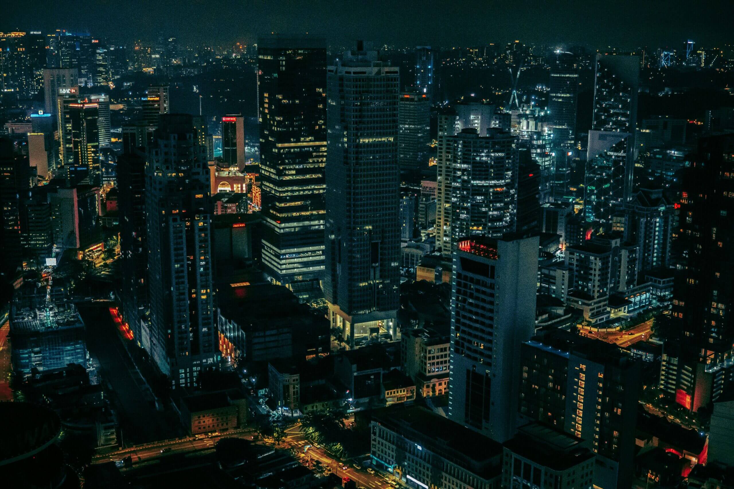 A nighttime aerial view of a bustling cityscape. Tall skyscrapers with illuminated windows dominate the scene, interspersed with smaller buildings and streets filled with car lights. The sky is dark, effortlessly setting the stage for the brightly lit urban environment. Rent reporting services keep this dynamic community thriving.
