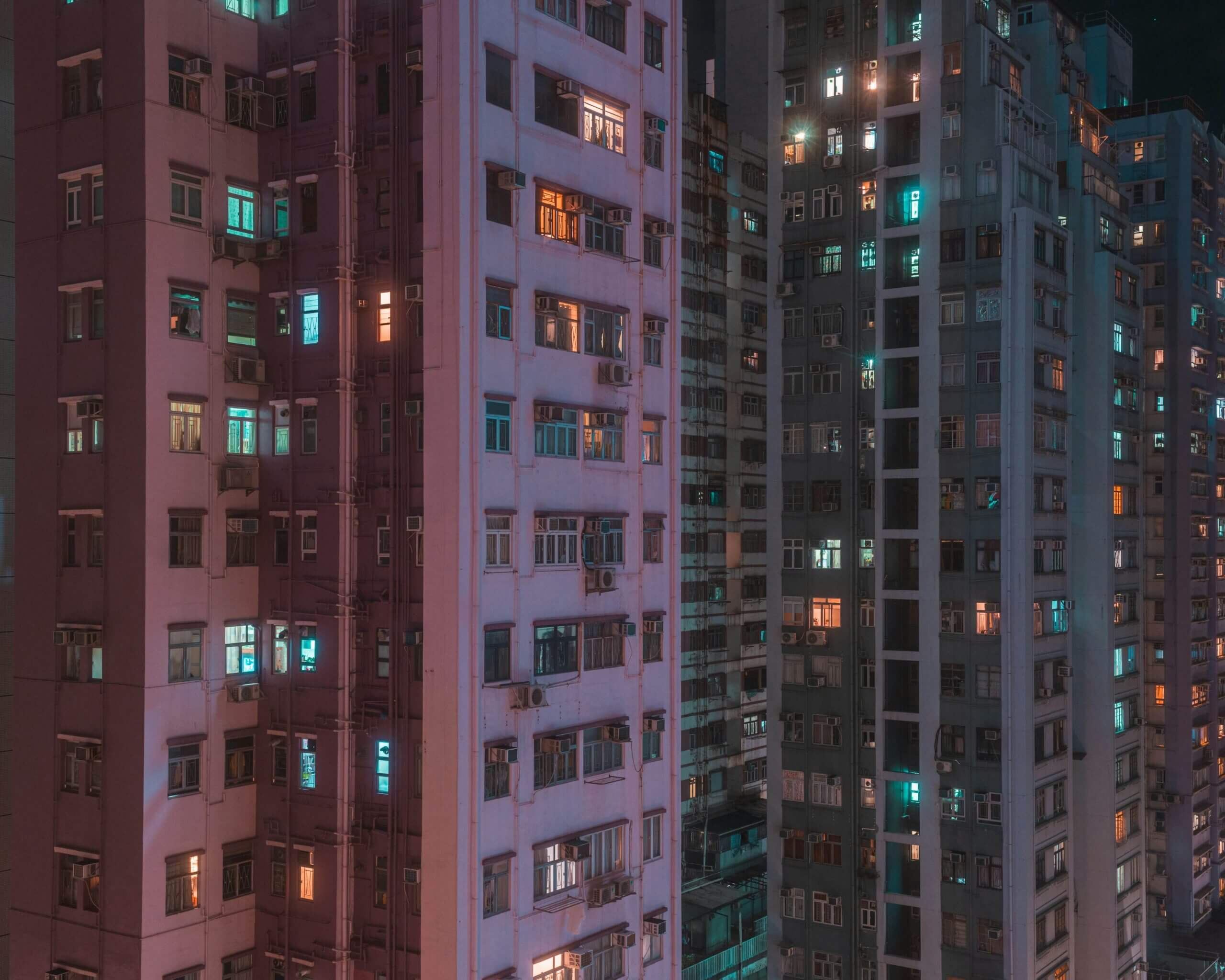 A night view of high-rise apartment buildings with colorful lights. Windows are illuminated in various hues of orange, blue, and white, creating a vibrant, urban atmosphere. The scene captures the density and height of city living and emphasizes the sense of community through rent reporting systems.