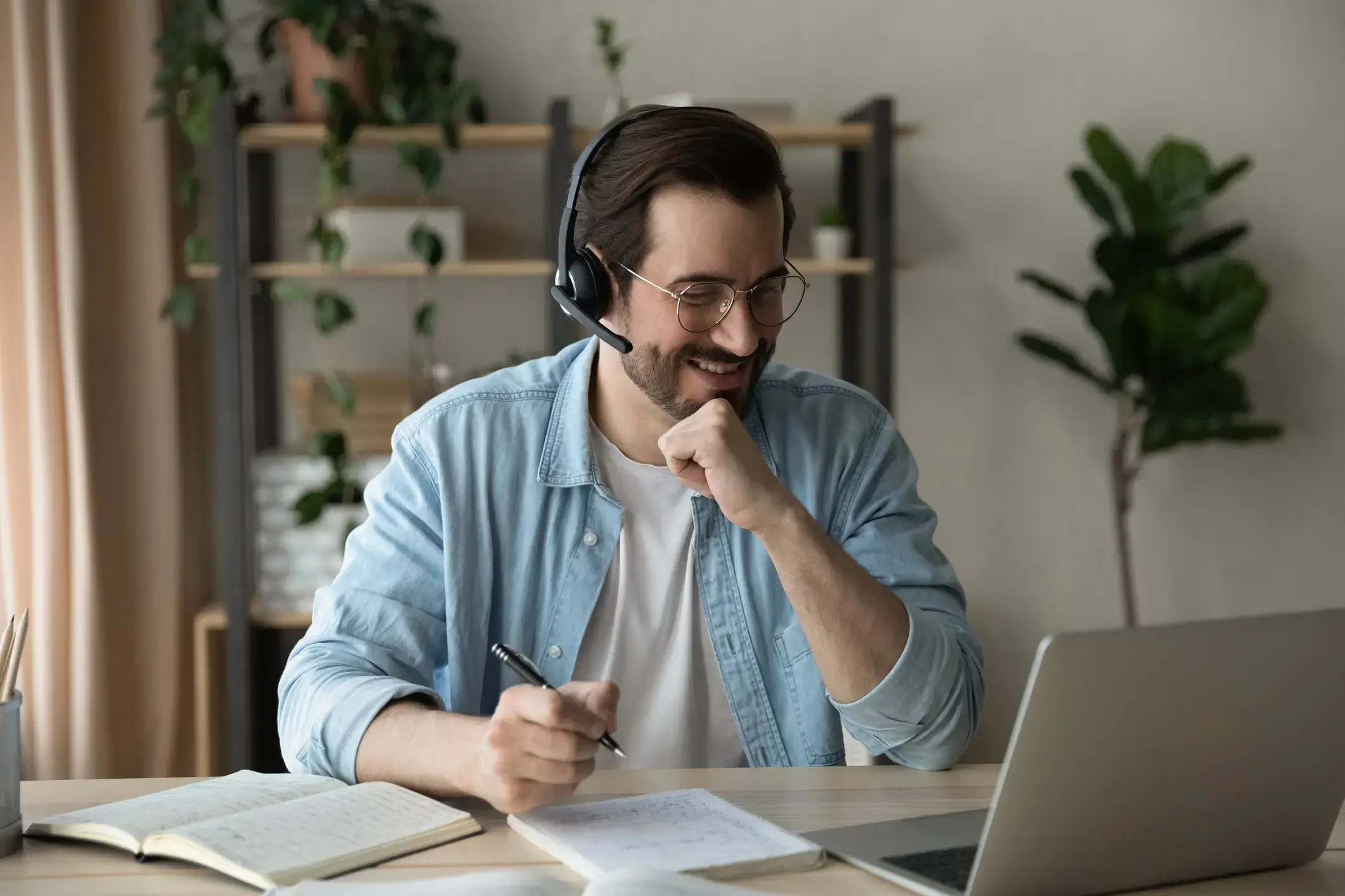 A man wearing glasses and a headset, smiling while looking at a laptop screen. He is seated at a desk with an open notebook, a pen in his hand, and a potted plant in the background. He appears to be engaged in an IDIQ video call or online meeting about credit management or rent reporting.