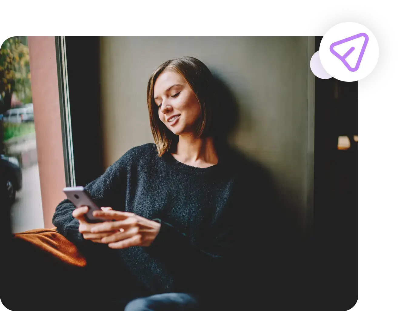 A person with short hair in a dark sweater sits by a window, smiling while looking at their phone. A chat icon with a purple outline is overlaid in the top right corner of the image. The scene subtly hints at discussions around employee benefits and identity theft protection.