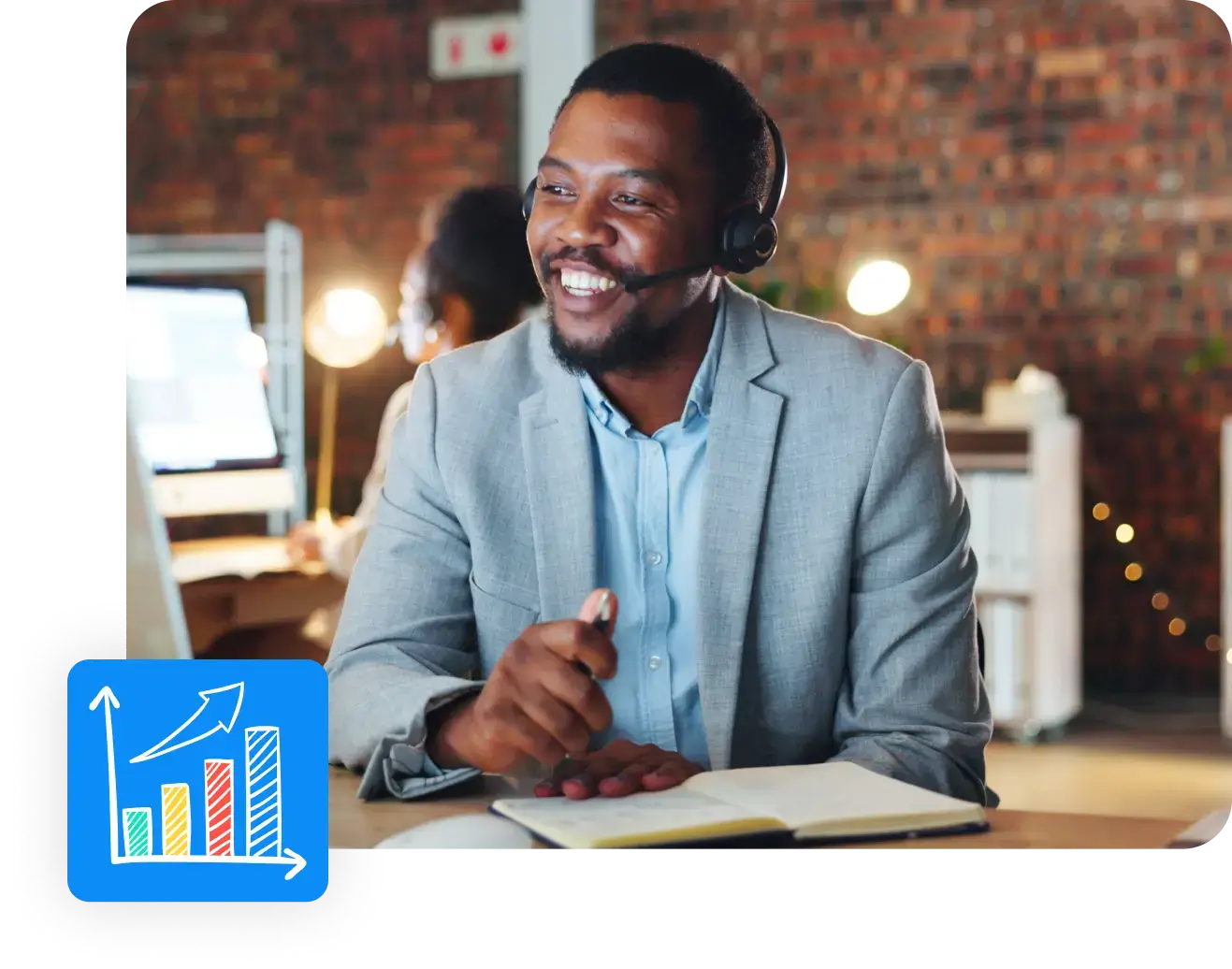 A smiling man in a gray suit and headset sits at a desk, gesturing with a pen while looking at a computer screen. An open notebook is in front of him. In the foreground, there's a graphic of a chart with upward bars and an arrow. The background shows a brick-walled office focused on employee benefits.