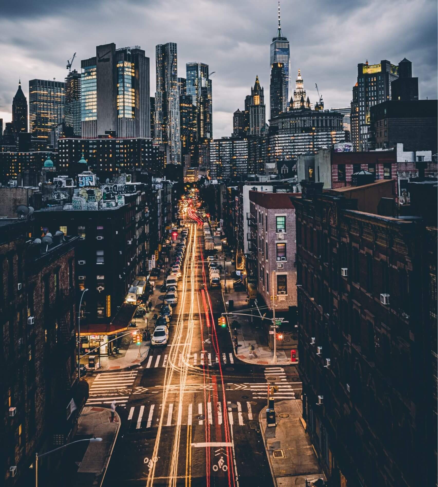 A bustling city street at dusk with long-exposure light trails from cars. Tall skyscrapers and buildings with illuminated windows dominate the background under a cloudy sky. The street, lined with lower residential buildings, boasts traffic lights and crosswalks, creating a scene where urban life meets employee benefits seamlessly.