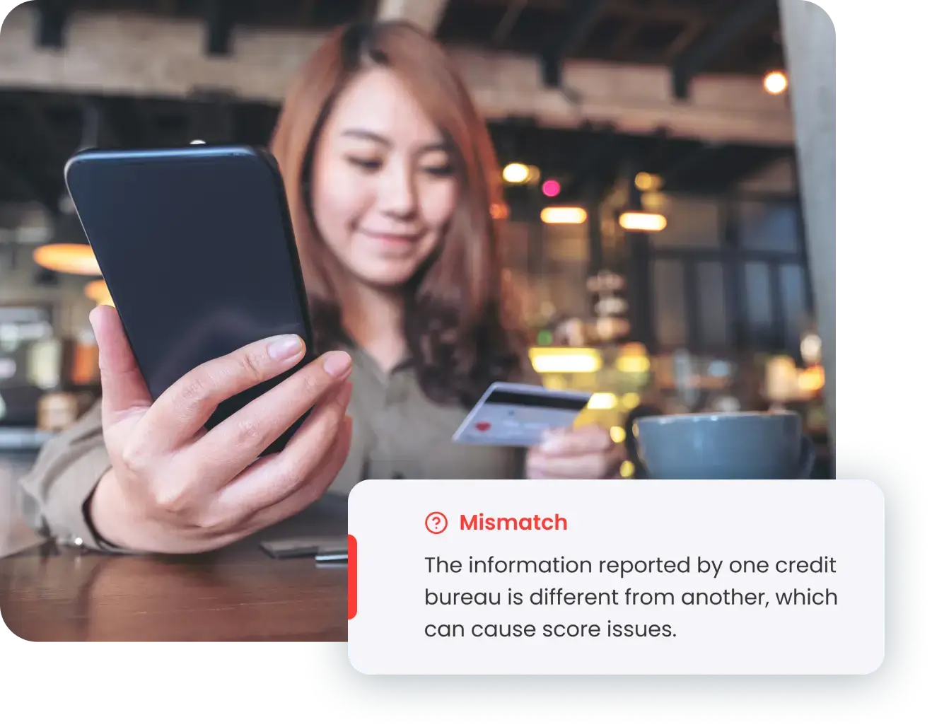 A woman with long hair is smiling and holding a smartphone in one hand and a credit card in the other, seated at a table in a warm, softly lit indoor setting. A notification about a credit report mismatch is displayed in the foreground, highlighting the importance of identity theft protection.