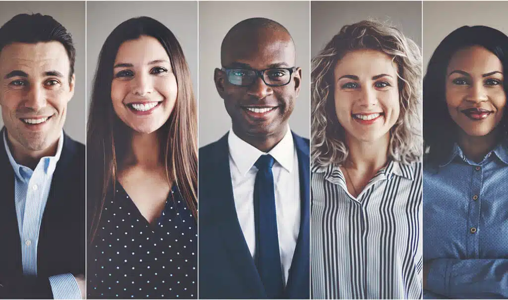 An image of a diverse group of five smiling professionals standing in a row. They are dressed in business attire and looking directly at the camera. Each individual appears confident and approachable, embodying the value of employee benefits. The background is a neutral, light-colored wall.