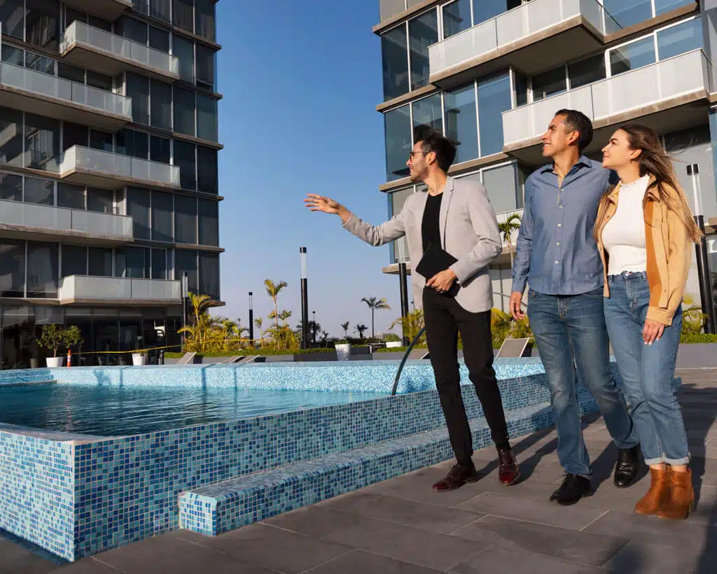 A real estate agent gestures towards a modern apartment building while showing it to a couple. They stand near a blue-tiled swimming pool situated between the apartment structures. All three individuals appear engaged and attentive. Palm trees are visible in the background, adding to the serene environment enhanced by identity theft protection features.