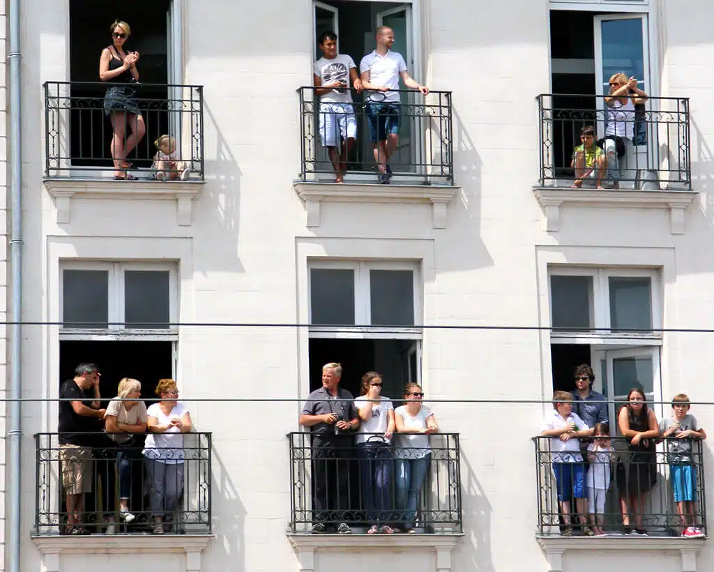People stand on small balconies of a white apartment building, enjoying the view. There are multiple groups, including children and adults, on the balconies. The scene suggests leisure and relaxation on a bright day while discussing rent reporting benefits.