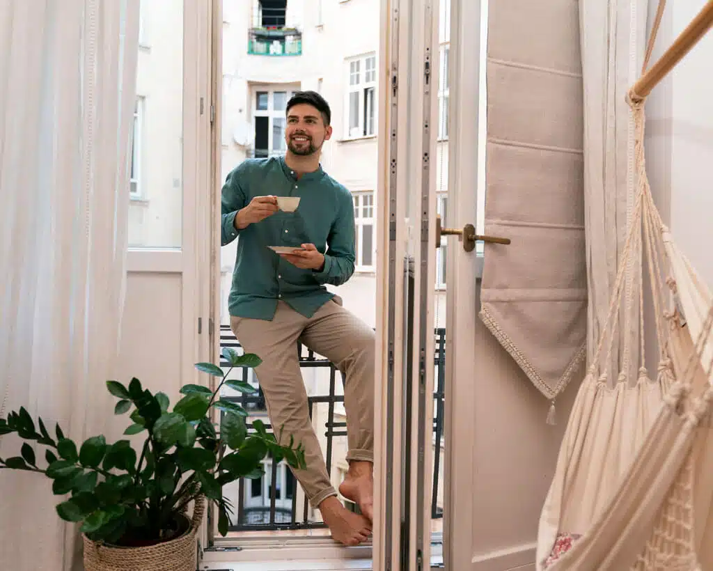 A man in a green shirt and beige pants relaxes with a cup of coffee on a balcony, leaning against the door frame. He's barefoot, and an indoor hammock and a potted plant are visible inside the cozy room. In the background are buildings and balconies, resembling scenes often associated with credit management ads.