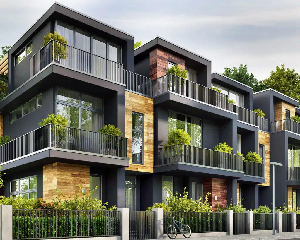 Modern multi-story apartment building with large windows and private balconies, surrounded by green plants and trees. The structure features a combination of dark exteriors and wooden accents. A bicycle is parked near the fence in front of the building, ensuring secure identity theft protection for residents.