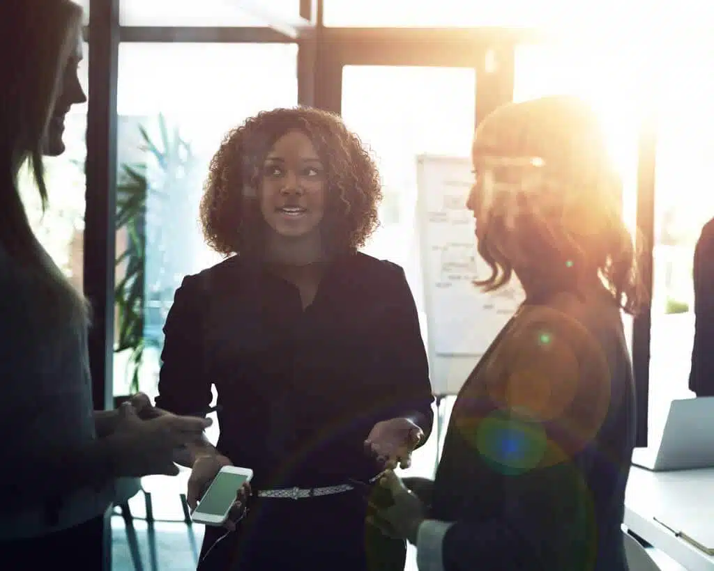 Three people stand in an office space, engaged in a conversation. The person in the center is holding a smartphone and looking at the coworker on the left. The bright sunlight streams in from the background, creating a warm and vibrant atmosphere as they discuss employee benefits.