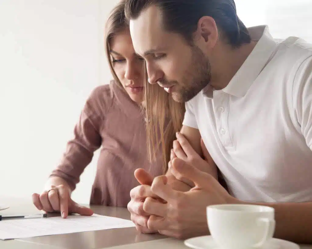 A man and woman sit closely together at a table, both focused on a document in front of them. The woman is pointing at the document. A cup of coffee and a pen are also on the table, suggesting they are reviewing paperwork related to employee benefits. The setting is bright and casual.