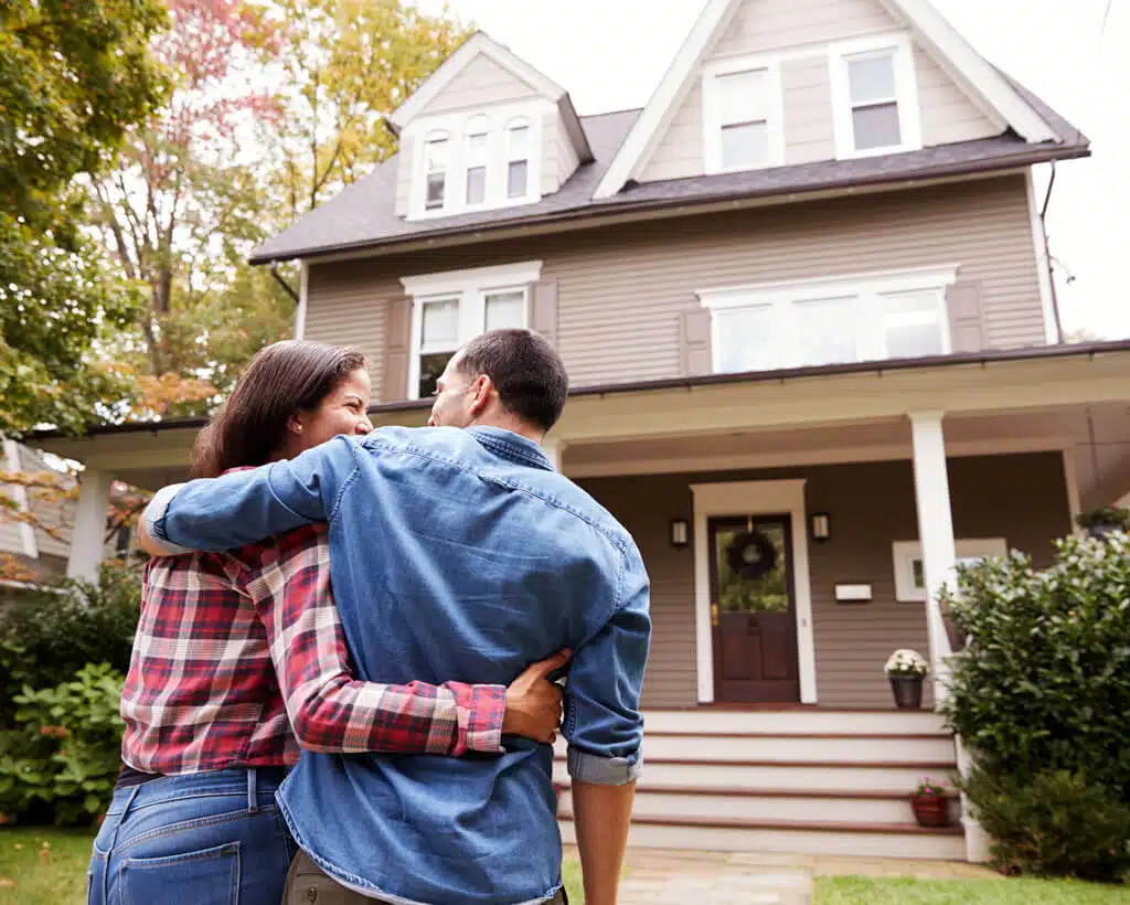 A couple stands closely together, arms around each other, facing a two-story house with a porch. The beige house with white trim is set amidst a green yard with trees. Both are wearing casual outfits; the woman in a plaid shirt and the man in a denim shirt. They recently invested in identity theft protection for peace of mind.