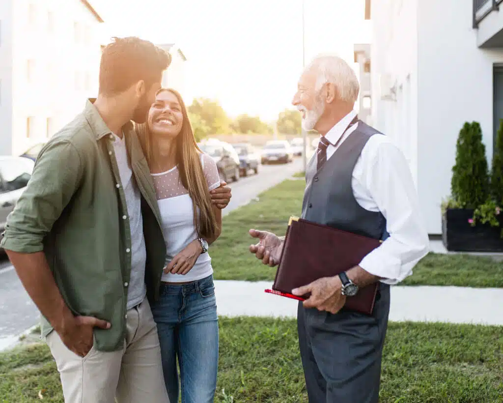A smiling couple is standing outside with an older man in a suit. The man is holding a folder and appears to be discussing rent reporting with the couple. They are standing on a sidewalk in front of houses, possibly talking about real estate options or identity theft protection.
