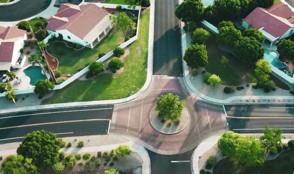 An aerial view of a suburban neighborhood intersection showing two roads meeting at a circular roundabout. The surrounding area features several houses with red-tiled roofs, green lawns, and swimming pools, all bordered by trees and neatly maintained landscaping—a testament to excellent credit management among residents.