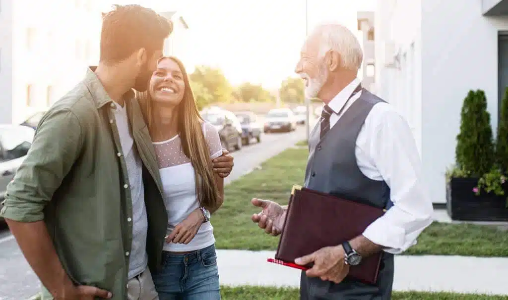A smiling couple stands outside with their arms around each other, talking to a senior man in a vest and tie who is holding a folder, possibly discussing credit management. They appear to be in a residential neighborhood on a sunny day.