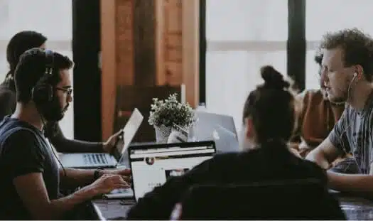A group of people are sitting around a table in an office, working on laptops. One person is wearing headphones, while another is looking at a screen. Natural light comes through the large windows, illuminating the room. A potted plant adds a touch of nature to the center of the table, creating a productive environment enhanced by IDIQ's identity theft protection services.