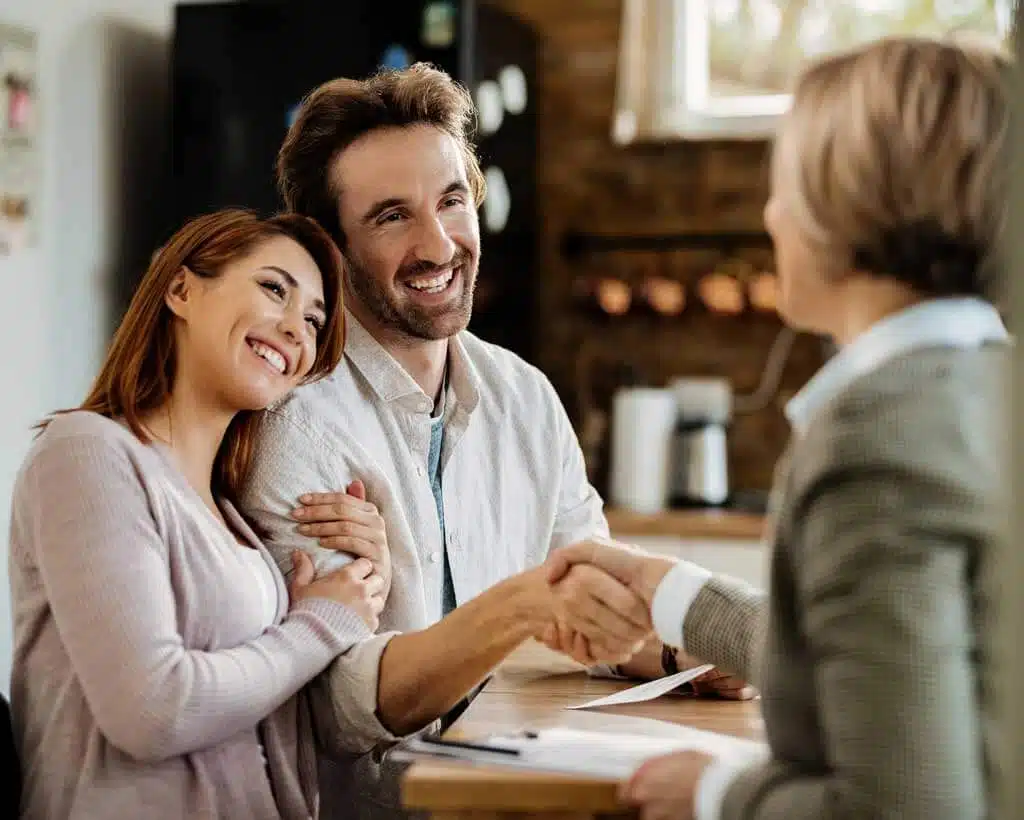 A happy couple sits at a table, smiling. The man is shaking hands with a woman opposite them, likely an advisor. The woman next to him leans in, looking content and supportive. They appear to be in a consultation about employee benefits or identity theft protection in an office setting.