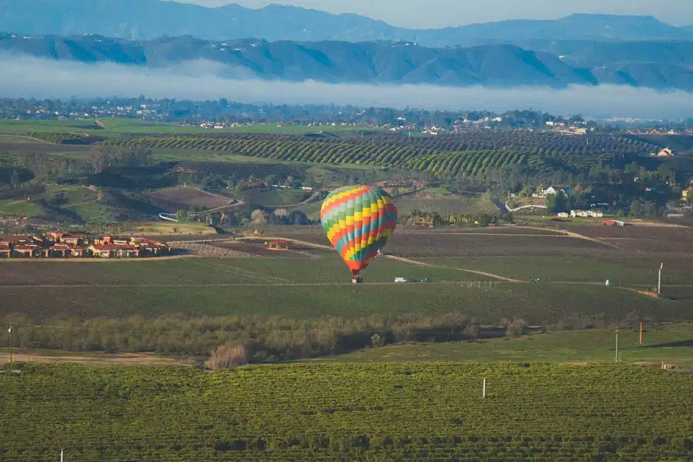 A multicolored hot air balloon floats low over a sprawling green landscape with fields and vineyards. In the background, hills and mountains are partially shrouded in fog under a clear blue sky. The scene is serene and expansive.