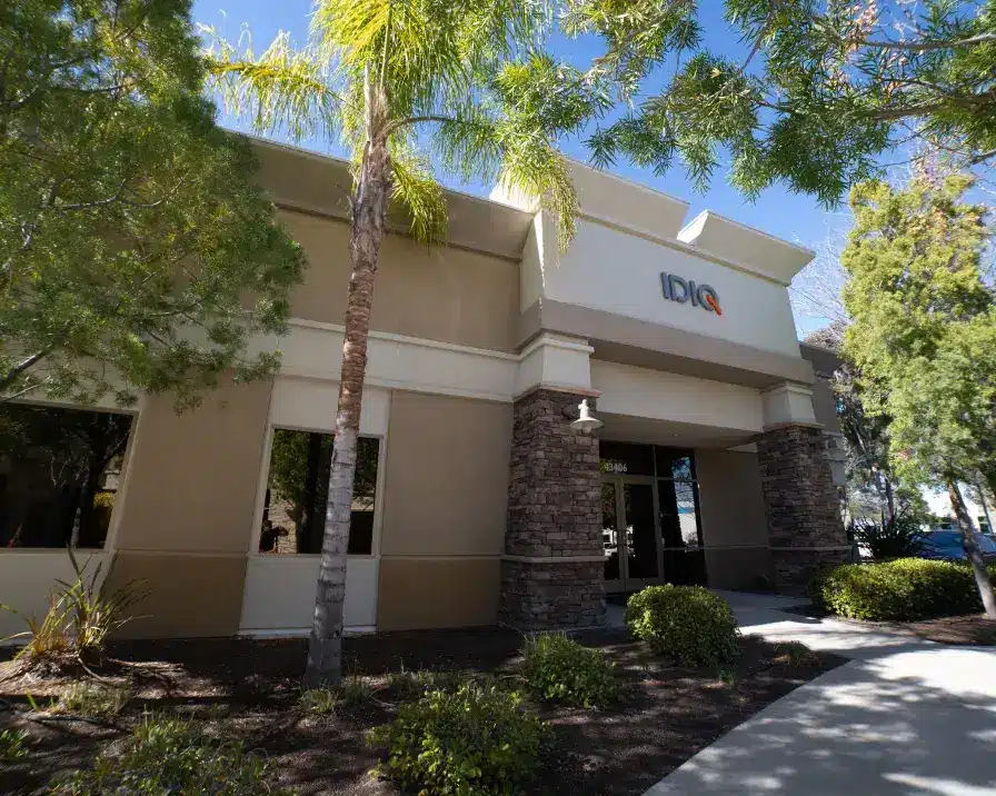 A modern office building is surrounded by palm trees and greenery. The building has a beige and cream facade with stone pillars flanking the entrance. The sign above the entrance reads "IDIQ," reflecting its services in credit management and identity theft protection. A sidewalk leads to the main doorway, with a clear sky overhead.