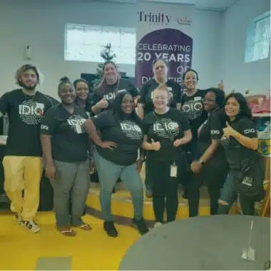 A group of ten smiling people stand together in a room, all wearing matching black T-shirts that say "ID10 GROUP". Behind them, a poster on the wall reads, "Trinity celebrating 20 years of digital impact." They pose with thumbs up and friendly gestures, promoting new employee benefits initiatives.