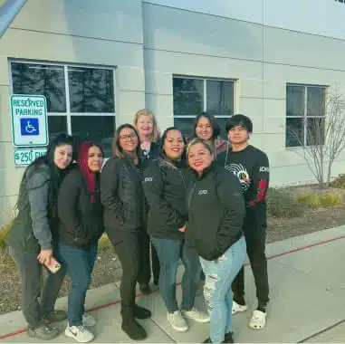 A group of eight people, including six women and two men, are standing together in front of a building. They are all wearing jackets and smiling at the camera. A sign indicating "Reserved Parking" for handicapped individuals is visible on the left. This moment showcases the importance of employee benefits for workplace satisfaction.