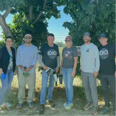 A group of six people stands outdoors under a tree on a sunny day. Some wear casual shirts, sunglasses, and caps, and two are wearing gloves, suggesting participation in a community or environmental activity. A chain-link fence and greenery are visible in the background, promoting awareness about rent reporting.