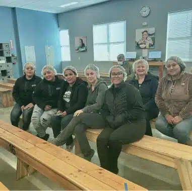 A group of seven people, wearing hair nets and casual clothing, sit on wooden benches in a well-lit room. Behind them are some windows, a wall clock, and two framed pictures. They all appear to be smiling and are positioned in a relaxed, informal setting, perhaps discussing employee benefits.